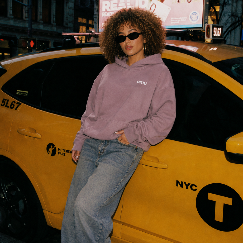 Person wearing a purple hoodie and sunglasses leaning against a yellow NYC taxi.